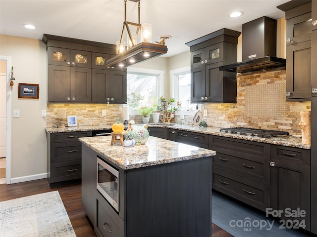 kitchen featuring dark wood-type flooring, wall chimney range hood, appliances with stainless steel finishes, decorative light fixtures, and a kitchen island