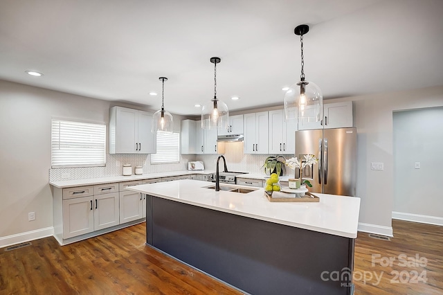 kitchen with backsplash, dark wood-type flooring, stainless steel refrigerator, hanging light fixtures, and an island with sink