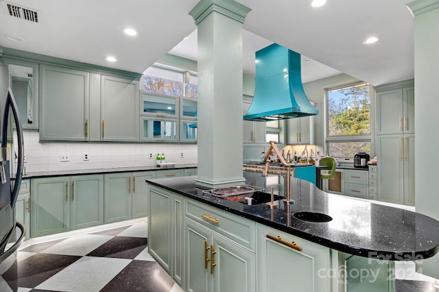 kitchen with island range hood, ornate columns, a kitchen island with sink, and dark stone counters