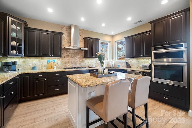 kitchen featuring tasteful backsplash, stainless steel appliances, wall chimney range hood, a center island, and a breakfast bar area