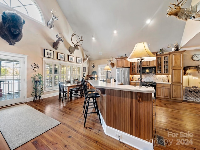 kitchen with black appliances, high vaulted ceiling, hardwood / wood-style floors, a breakfast bar area, and an island with sink