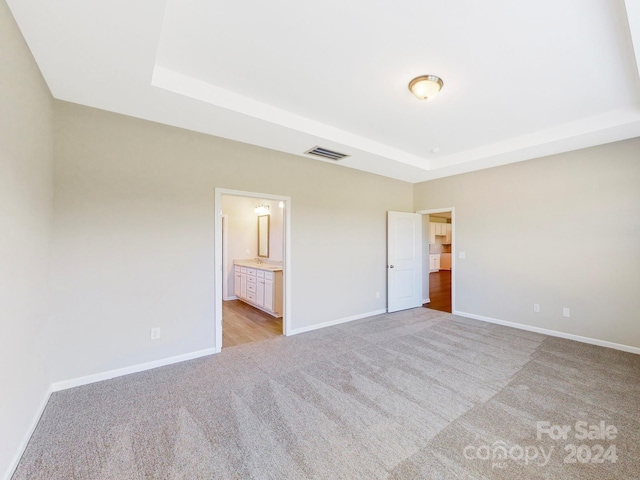 unfurnished bedroom featuring a raised ceiling, light colored carpet, and ensuite bath