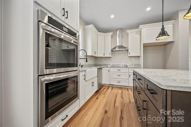 kitchen featuring light wood finished floors, appliances with stainless steel finishes, white cabinetry, wall chimney exhaust hood, and a sink