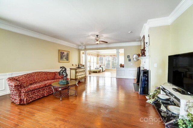 living area with a ceiling fan, wood finished floors, wainscoting, crown molding, and decorative columns