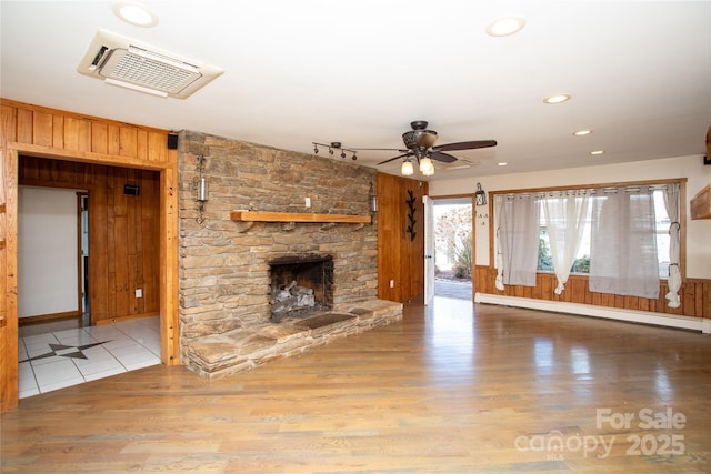 unfurnished living room with track lighting, ceiling fan, wooden walls, a baseboard heating unit, and a stone fireplace