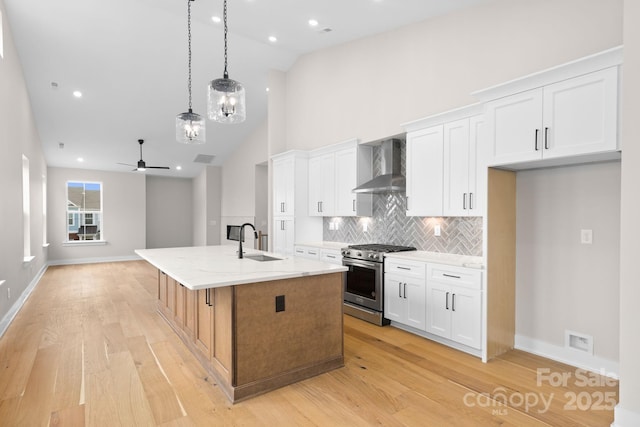 kitchen featuring wall chimney range hood, light stone counters, stainless steel range, an island with sink, and white cabinets