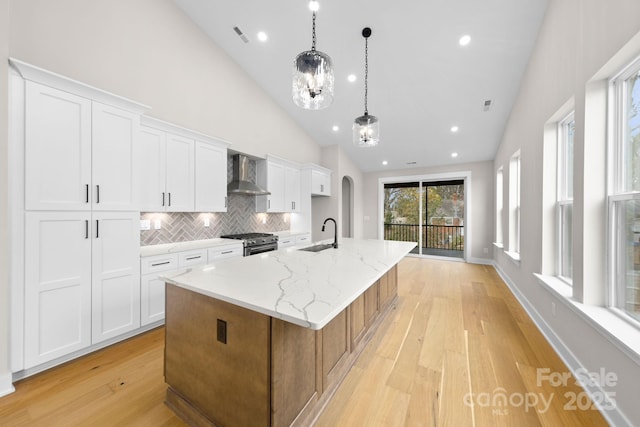 kitchen with stainless steel stove, hanging light fixtures, white cabinets, an island with sink, and wall chimney exhaust hood