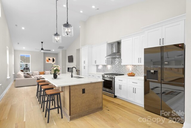 kitchen featuring stainless steel range, black fridge with ice dispenser, white cabinetry, wall chimney range hood, and an island with sink