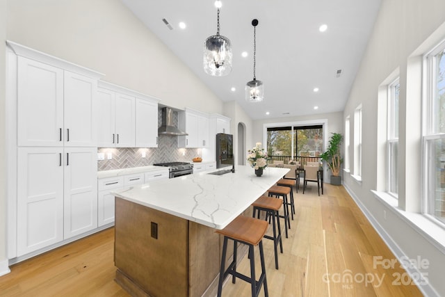 kitchen with a center island with sink, white cabinets, wall chimney exhaust hood, hanging light fixtures, and high end stove