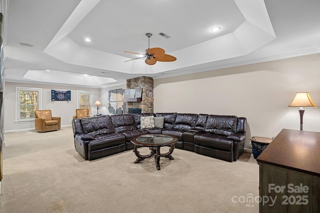 carpeted living room with a tray ceiling, a stone fireplace, ceiling fan, and ornamental molding