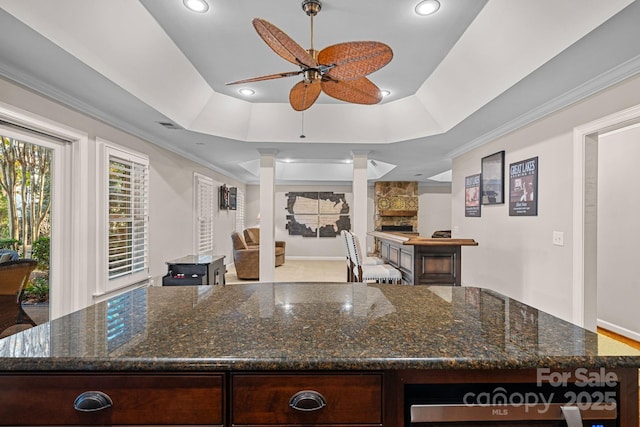 kitchen featuring a tray ceiling, a stone fireplace, ceiling fan, and dark stone counters