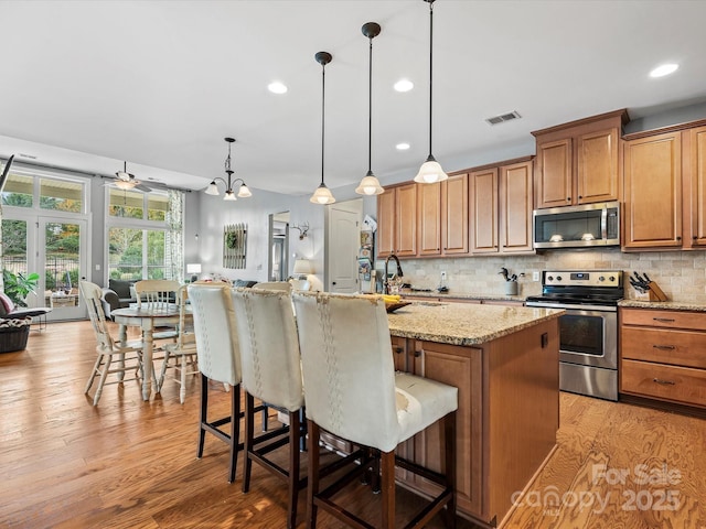 kitchen with ceiling fan with notable chandelier, hanging light fixtures, light stone countertops, an island with sink, and stainless steel appliances