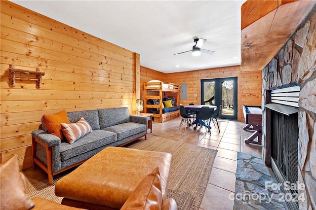 tiled living room featuring french doors, a stone fireplace, ceiling fan, and wood walls