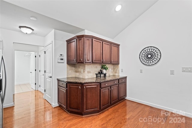 kitchen with decorative backsplash, stainless steel fridge, dark stone counters, vaulted ceiling, and light hardwood / wood-style flooring