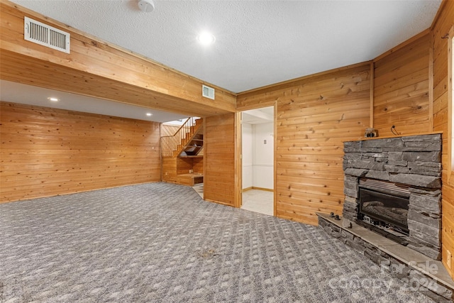 unfurnished living room with a stone fireplace, wooden walls, carpet floors, and a textured ceiling