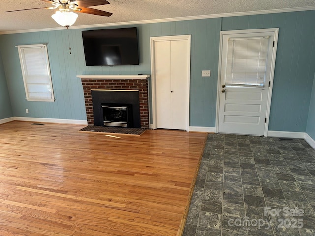 unfurnished living room with wood walls, a brick fireplace, ceiling fan, ornamental molding, and a textured ceiling