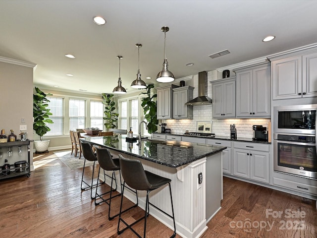 kitchen featuring gray cabinetry, wall chimney exhaust hood, dark stone counters, a kitchen bar, and ornamental molding