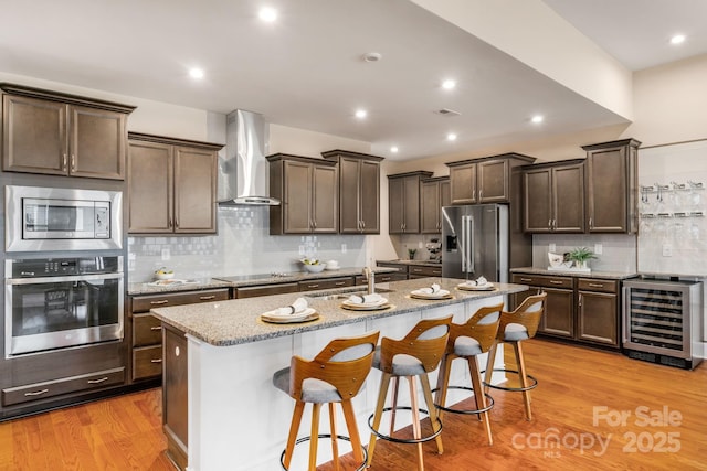 kitchen with wall chimney exhaust hood, wine cooler, an island with sink, a breakfast bar area, and appliances with stainless steel finishes