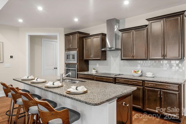 kitchen featuring appliances with stainless steel finishes, hardwood / wood-style flooring, a center island with sink, and wall chimney range hood