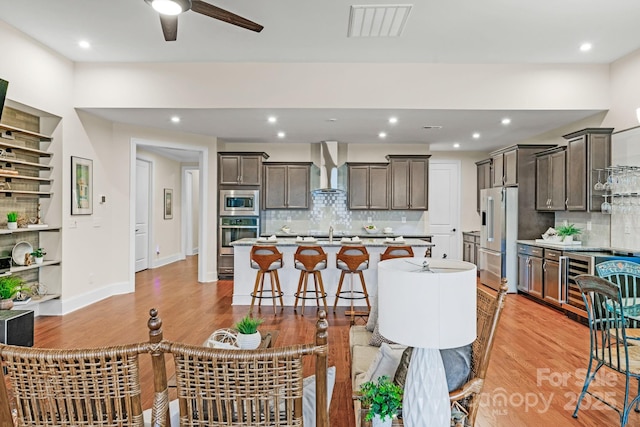 kitchen featuring backsplash, a center island with sink, wall chimney exhaust hood, light hardwood / wood-style floors, and stainless steel appliances