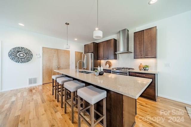 kitchen featuring appliances with stainless steel finishes, an island with sink, light hardwood / wood-style floors, and wall chimney range hood