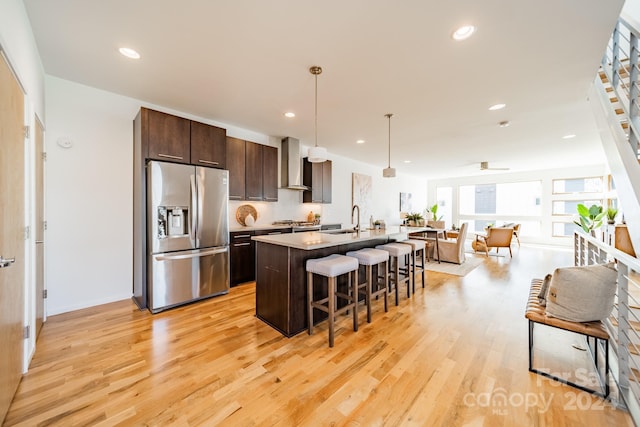 kitchen with stainless steel appliances, wall chimney range hood, decorative light fixtures, a kitchen island with sink, and a breakfast bar