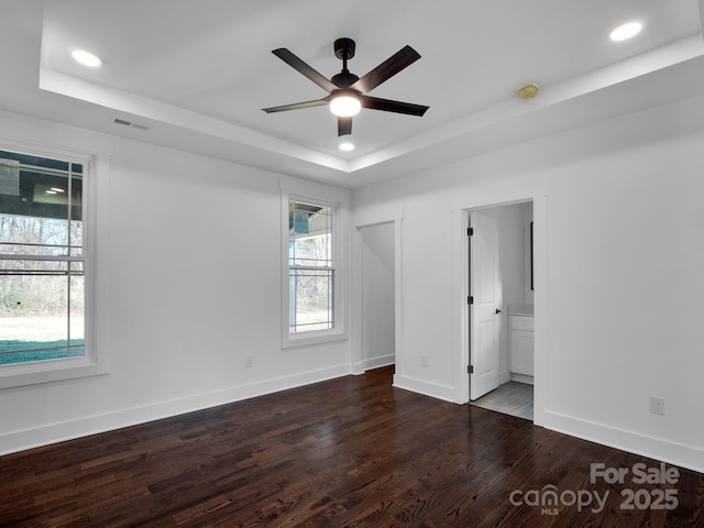 unfurnished bedroom featuring ensuite bath, a raised ceiling, ceiling fan, multiple windows, and dark hardwood / wood-style floors