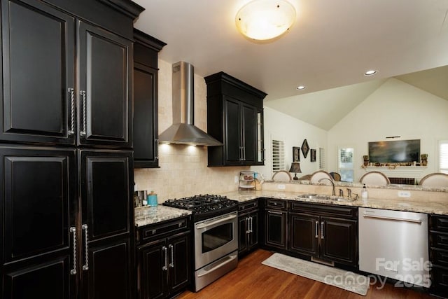 kitchen with light stone counters, stainless steel appliances, dark cabinetry, wall chimney range hood, and a sink