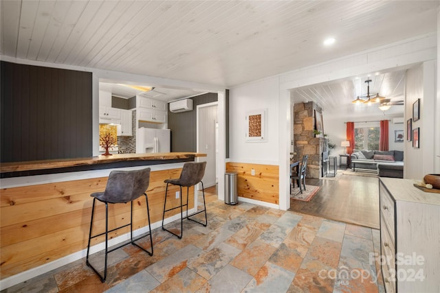 kitchen featuring wooden walls, an AC wall unit, white cabinetry, white fridge with ice dispenser, and a breakfast bar area