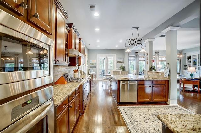 kitchen featuring ornate columns, appliances with stainless steel finishes, backsplash, hanging light fixtures, and light stone counters