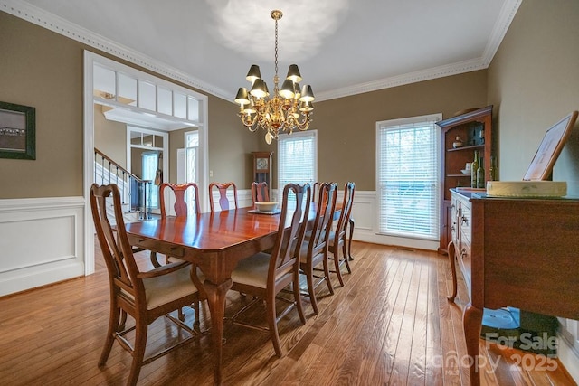 dining room with a wainscoted wall, crown molding, light wood-style flooring, a chandelier, and stairs