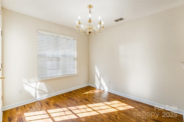 empty room with visible vents, an inviting chandelier, a textured ceiling, wood finished floors, and baseboards