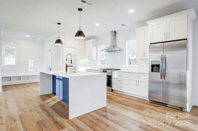 kitchen with a kitchen island with sink, white cabinets, stainless steel appliances, and wall chimney range hood