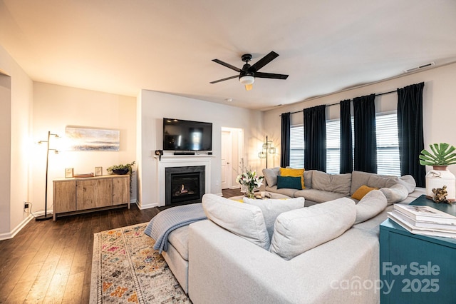 living area featuring visible vents, baseboards, a glass covered fireplace, dark wood-style floors, and ceiling fan