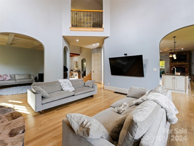 living room featuring beam ceiling, ceiling fan, a brick fireplace, a towering ceiling, and light hardwood / wood-style floors