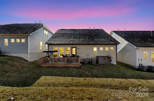 back house at dusk with a deck, central AC unit, and a lawn