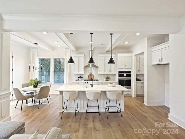 kitchen featuring white cabinets, an island with sink, double oven, decorative light fixtures, and light hardwood / wood-style floors