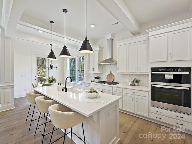 kitchen with wall chimney range hood, sink, an island with sink, light hardwood / wood-style floors, and white cabinetry