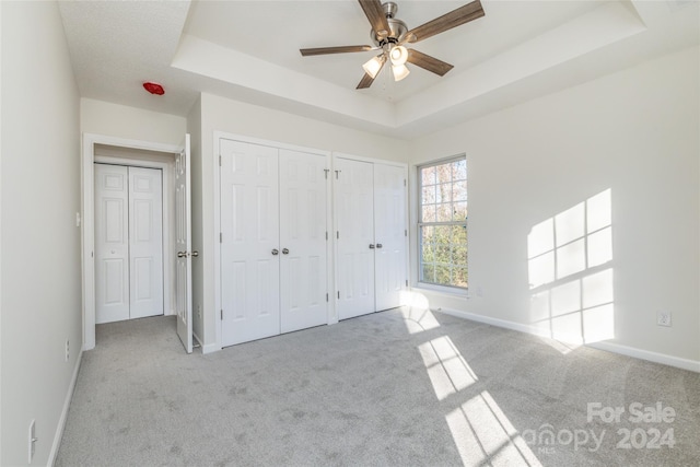 unfurnished bedroom featuring a raised ceiling, multiple closets, ceiling fan, and light carpet