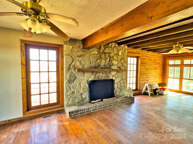 unfurnished living room with beam ceiling, ceiling fan, a stone fireplace, hardwood / wood-style floors, and a textured ceiling