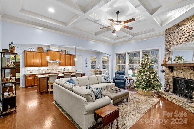 living room with ornamental molding, coffered ceiling, dark wood-type flooring, beamed ceiling, and a stone fireplace