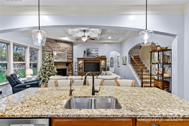 kitchen with sink, coffered ceiling, a stone fireplace, light stone counters, and pendant lighting