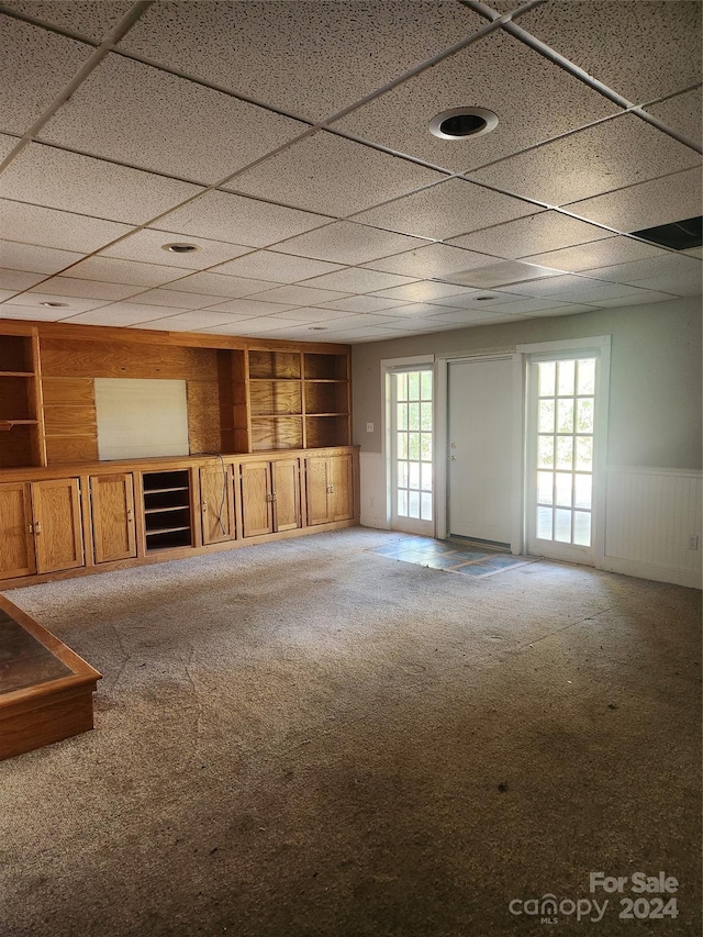 unfurnished living room with carpet flooring, a drop ceiling, wood walls, and built in features