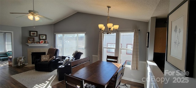 dining area featuring ceiling fan with notable chandelier, lofted ceiling, a tiled fireplace, dark wood-type flooring, and a textured ceiling