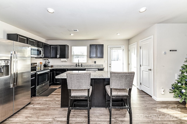 kitchen featuring appliances with stainless steel finishes, a kitchen island, light stone counters, wood-type flooring, and a breakfast bar area