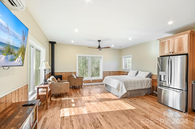 bedroom featuring a wood stove, an AC wall unit, stainless steel refrigerator with ice dispenser, ceiling fan, and light wood-type flooring