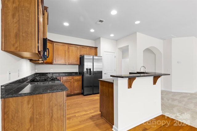 kitchen featuring black gas stovetop, a kitchen island with sink, light hardwood / wood-style flooring, dark stone countertops, and stainless steel fridge with ice dispenser