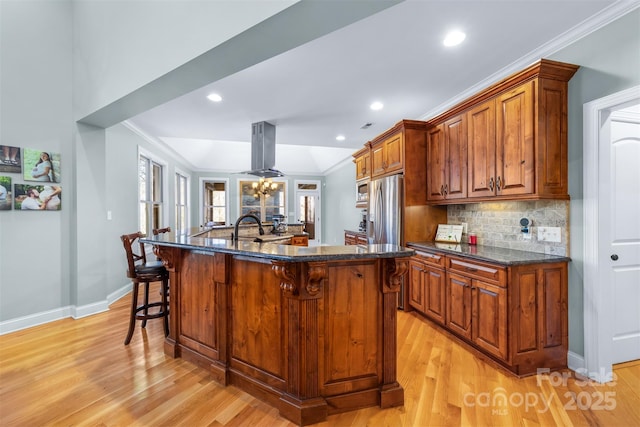 kitchen with island range hood, dark stone counters, ornamental molding, stainless steel appliances, and a kitchen island with sink