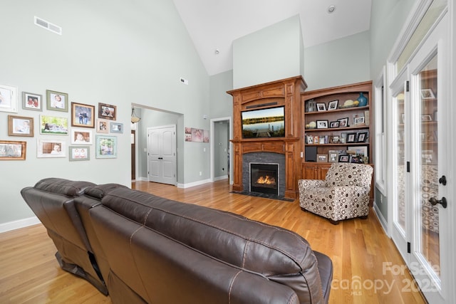 living area with high vaulted ceiling, light wood finished floors, a fireplace with flush hearth, baseboards, and visible vents