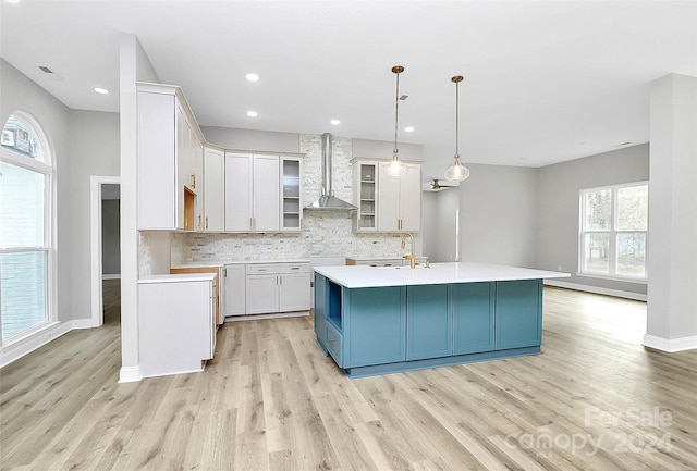 kitchen featuring blue cabinetry, wall chimney exhaust hood, hanging light fixtures, a kitchen island with sink, and light wood-type flooring